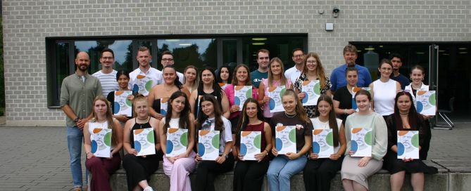 Gruppenfoto von Absolventinnen und Absolventen mit ihren Abschlusszeugnissen vor einem Schulgebäude. In der ersten Reihe sitzen junge Frauen mit Zeugnismappen auf dem Schoß, dahinter stehen weitere junge Menschen sowie Lehrkräfte. Alle lächeln in die Kamera.