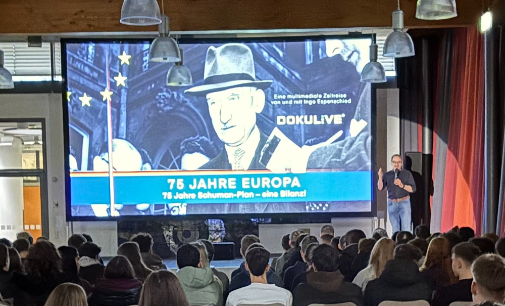 Referent hält einen Vortrag vor vielen Jugendlichen in einer Mensa; auf der Leinwand steht „75 Jahre Europa – 75 Jahre Schuman-Plan