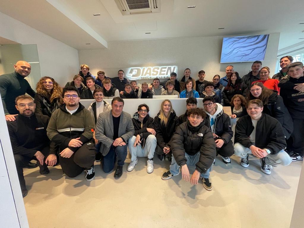 Große Gruppe von Schülern und Erwachsenen steht gemeinsam in einem Unternehmensgebäude vor einem Firmenlogo.