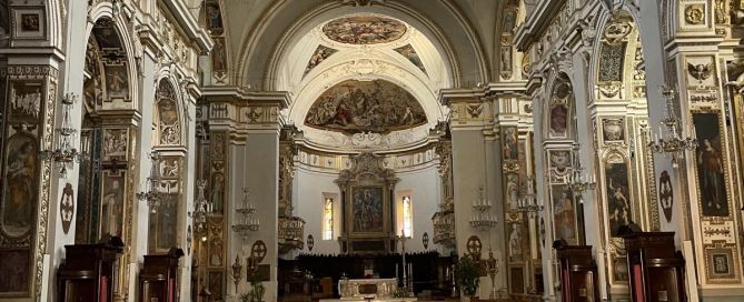 Blick durch den Innenraum einer prächtig ausgestatteten Kirche in Fabriano. Der lange Mittelgang mit schwarz-weißem Schachbrettboden führt zum Altar. Auf beiden Seiten stehen hölzerne Kirchenbänke. Kunstvolle Wandmalereien und verzierte Säulen prägen den Kirchenraum.