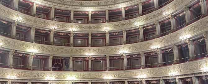 Blick in einen historischen italienischen Theatersaal mit mehreren übereinanderliegenden Logenreihen. Goldene Verzierungen, rote Samtbezüge und eine aufwendig bemalte Decke mit Kronleuchter prägen die Architektur.