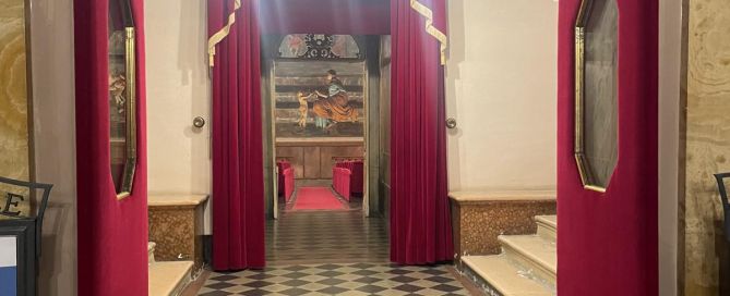 Blick durch geöffnete rote Schwingtüren in den Eingangsbereich eines historischen Theaters in Fabriano. Rote Samtvorhänge, ein Kronleuchter und ein schwarz-weiß gemusterter Fliesenboden prägen den Raum.